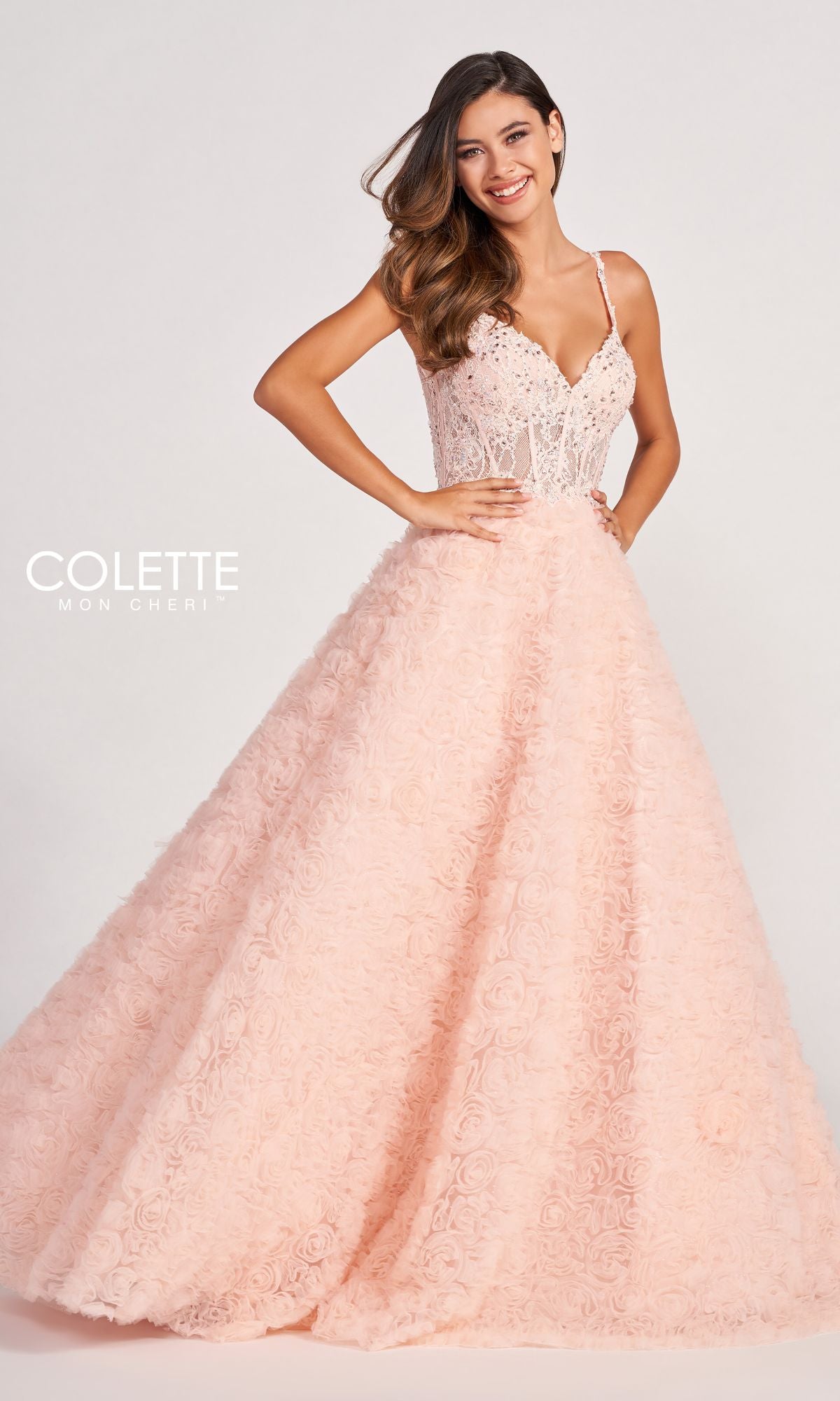 A woman stands against a plain background, smiling in the Colette Sheer-Bodice Rosette Beaded Lace Ball Gown featuring a fitted embellished bodice and voluminous textured skirt in light pink.